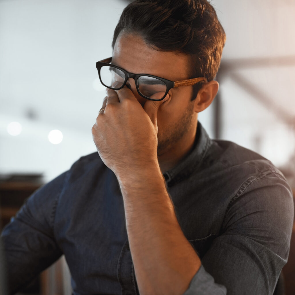 Man under Stress And Eye Strain In Office Stressed man in glasses rubbing eyes, possibly tired or overworked, sitting at a desk in a dimly lit room.