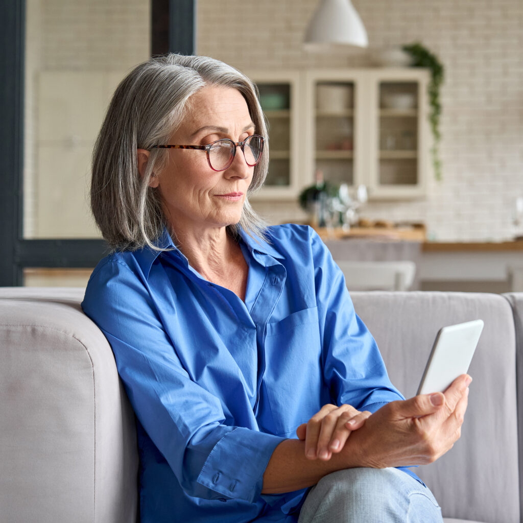 Serious Mature Middle Age Senior Woman At Home,On Couch Senior woman in a blue shirt using a smartphone on a couch at home, looking thoughtful.
