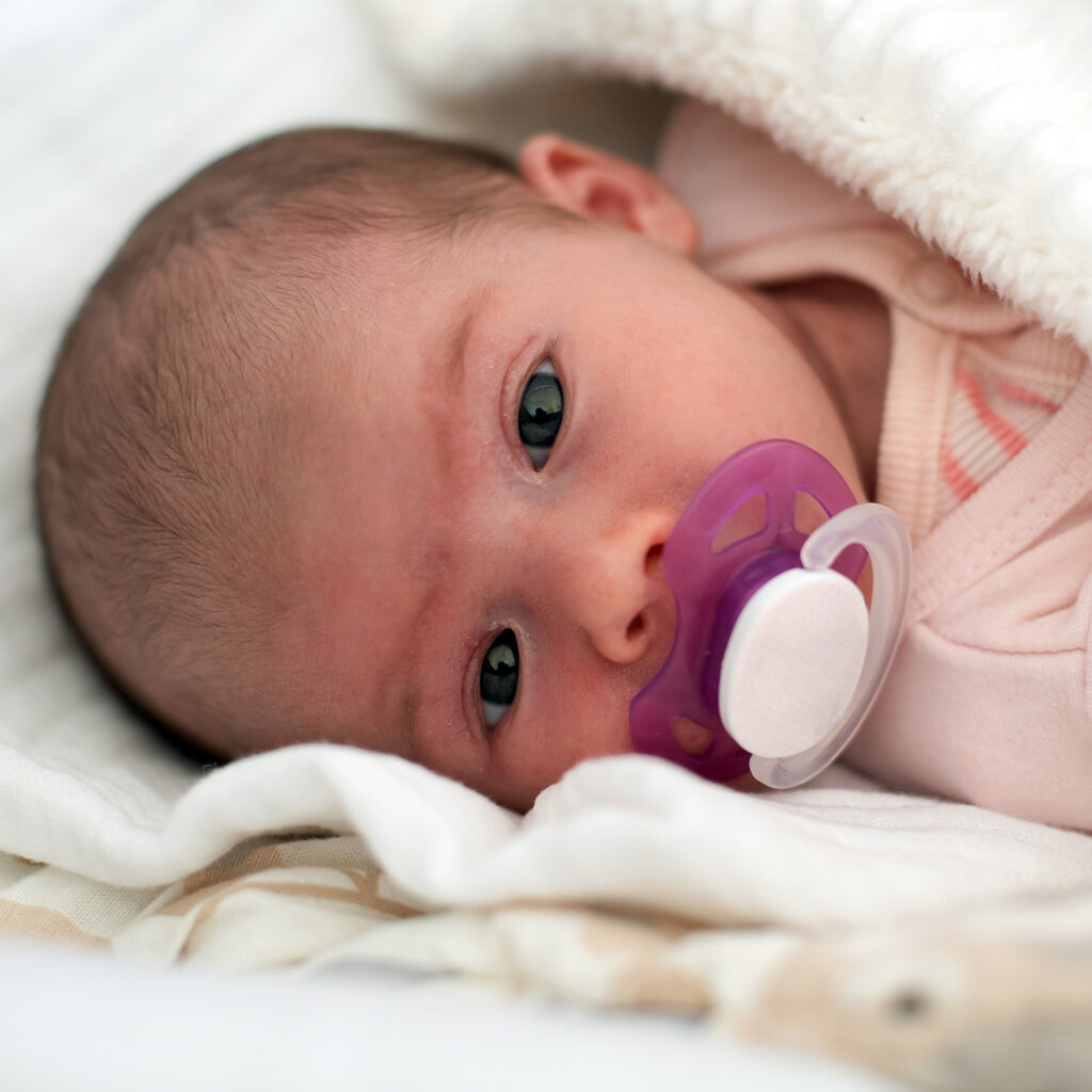 Newborn Baby Portrait Newborn baby with purple pacifier lying on a soft white blanket, looking calm and cozy.