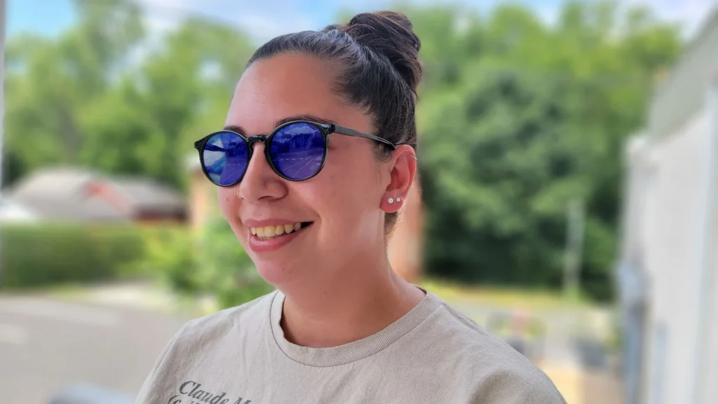 Smiling person wearing sunglasses with blue lenses outside on a sunny day.