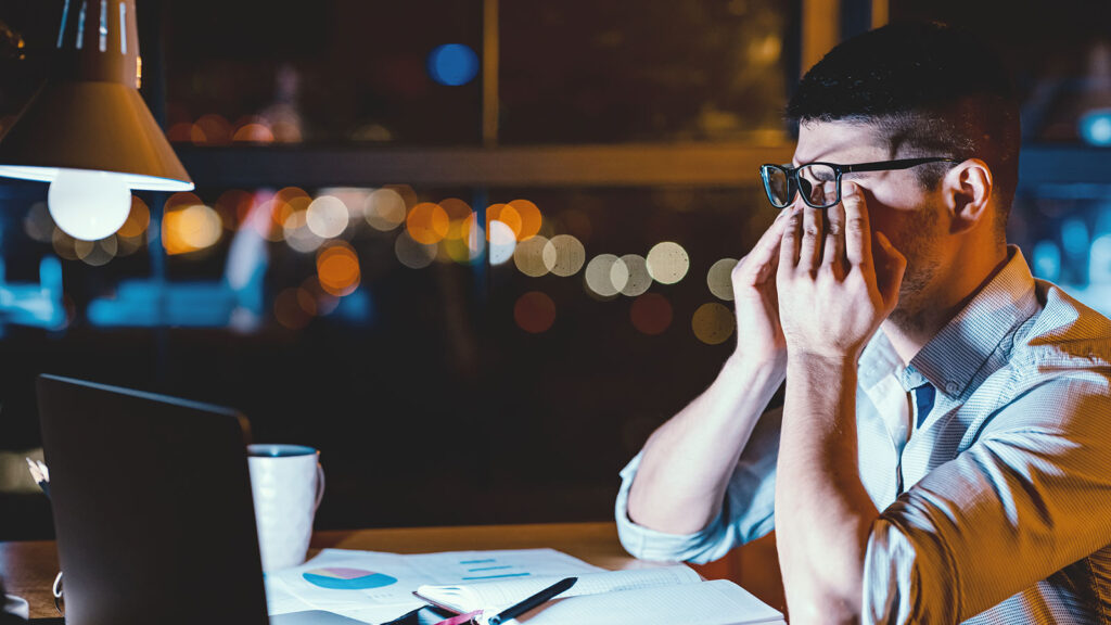 Man in glasses rubs eyes while working late on laptop, surrounded by papers and coffee, illuminated by desk lamp.