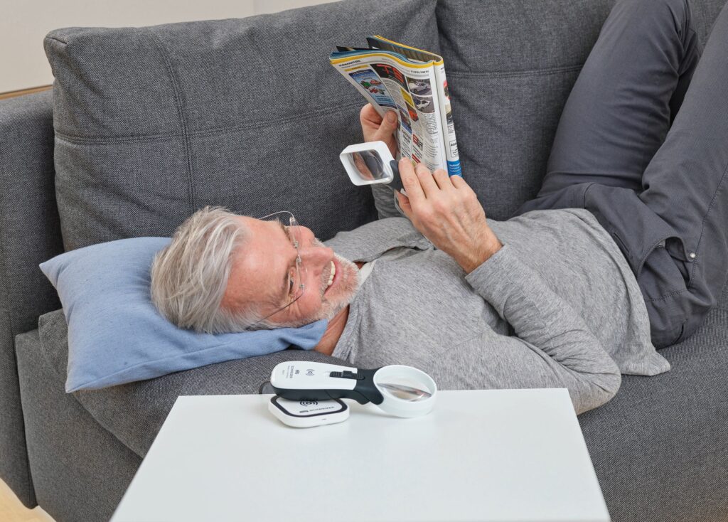 Man relaxing on a couch, reading a magazine with magnifier glasses, next to a table with magnifying tools.