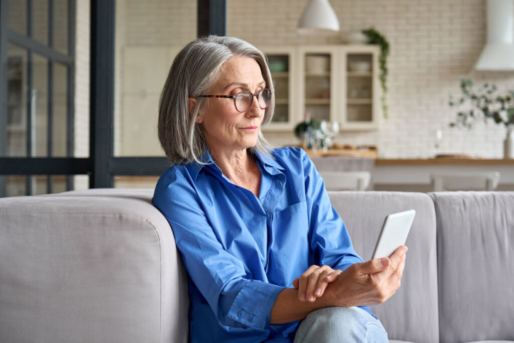 Older woman in blue shirt using smartphone on couch at home.