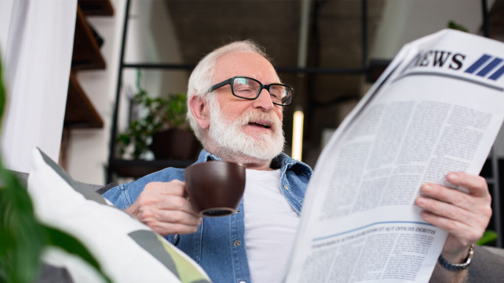 Elderly man enjoying a coffee while reading a newspaper at home, surrounded by greenery.