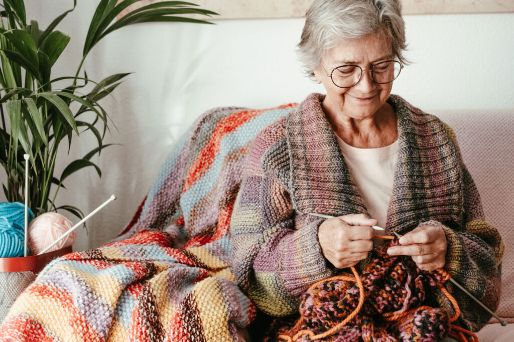 Elderly woman knitting colorful blanket, sitting on sofa, plants and yarn beside her. Cozy and creative atmosphere.