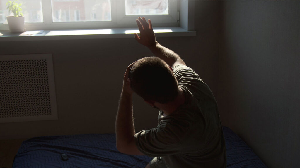Person shielding eyes from sunlight near a window, sitting on a bed with a blue coverlet.