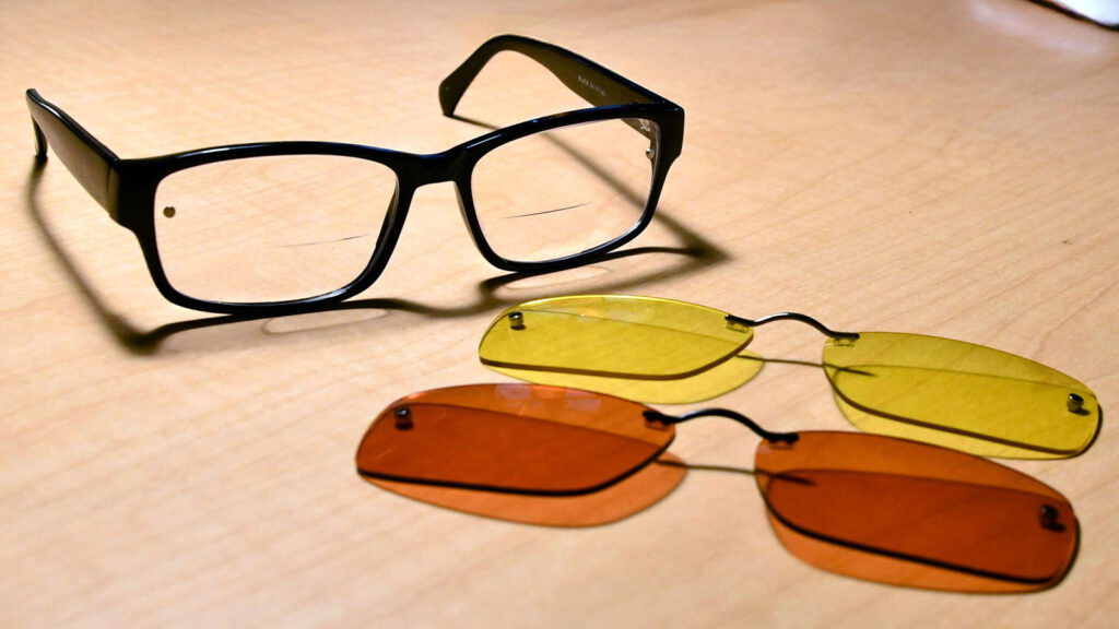 Black eyeglasses with interchangeable yellow and orange lenses on a wooden surface.