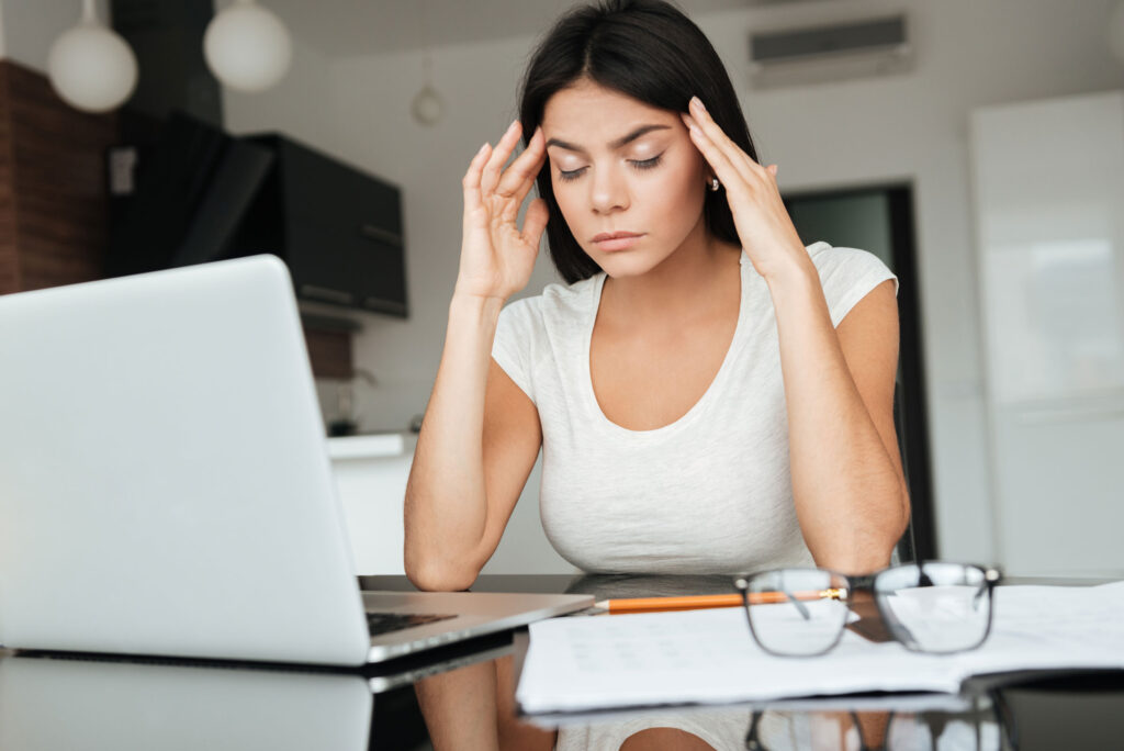 Woman feeling stressed at work, sitting at a desk with a laptop, holding her head in her hands.