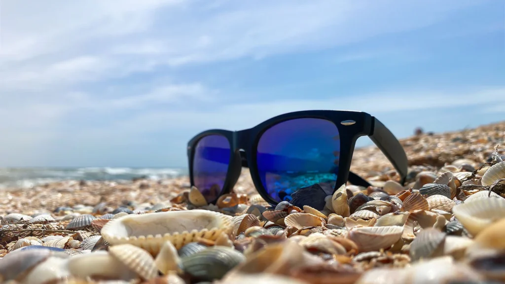 Sunglasses on a shell-covered beach under a clear sky, reflecting the ocean waves.