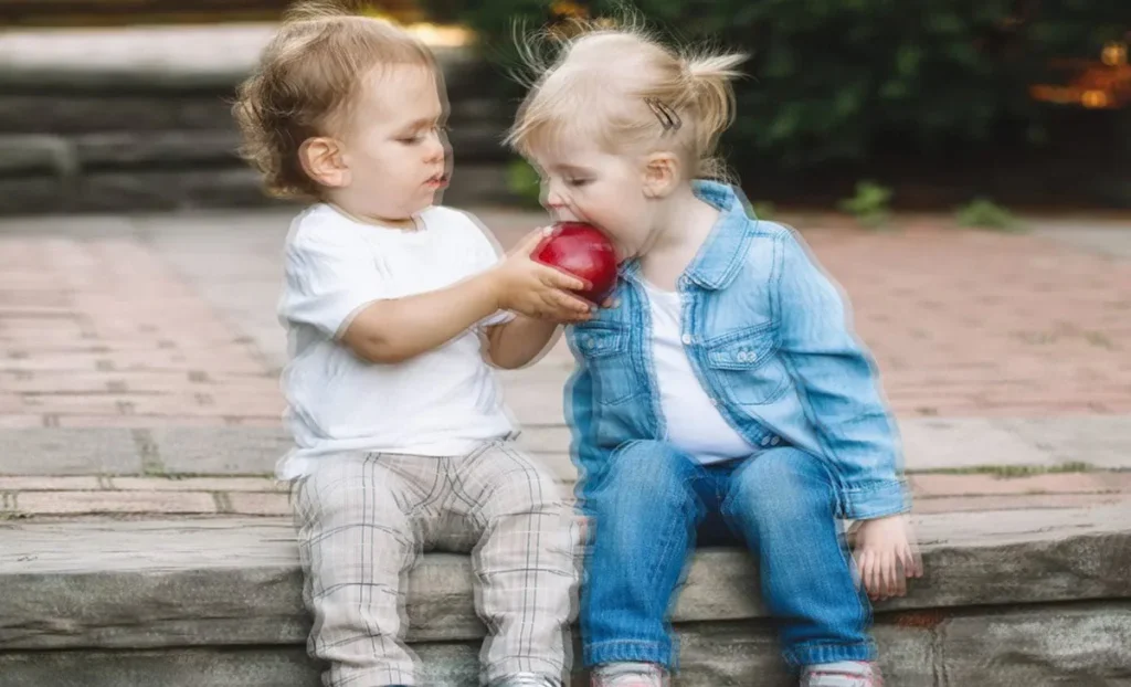 Two toddlers sharing a red apple while sitting on a stone step outdoors.
