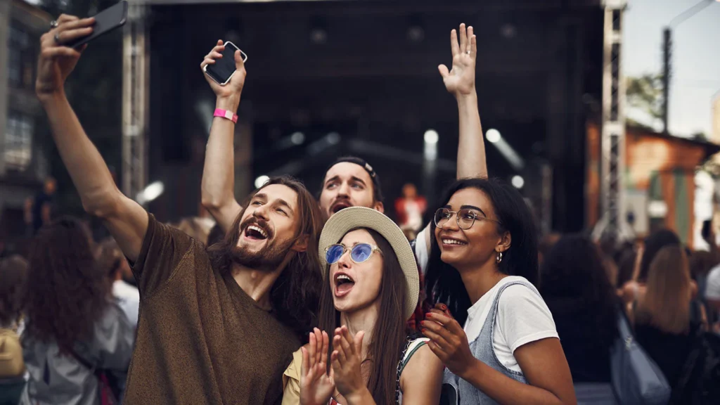Group of friends enjoying a festival, taking selfies and cheering in front of a live stage.