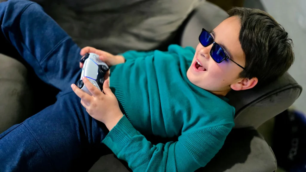 Child wearing sunglasses happily playing video games on a couch with a controller.