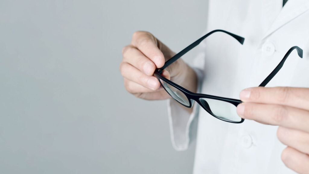 Person in white coat holding a pair of black eyeglasses, gray background.