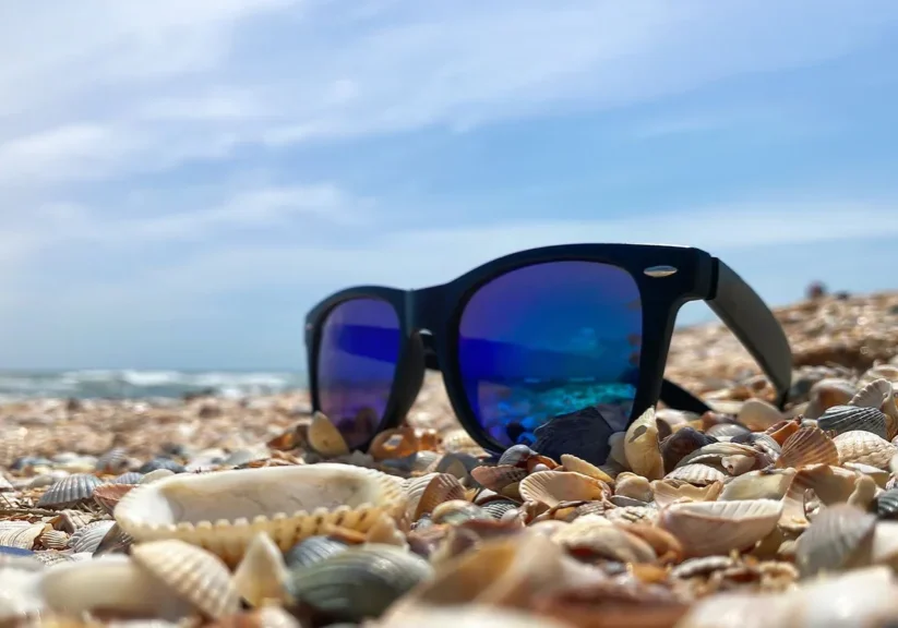 Sunglasses on a shell-covered beach under a clear sky, reflecting the ocean waves.