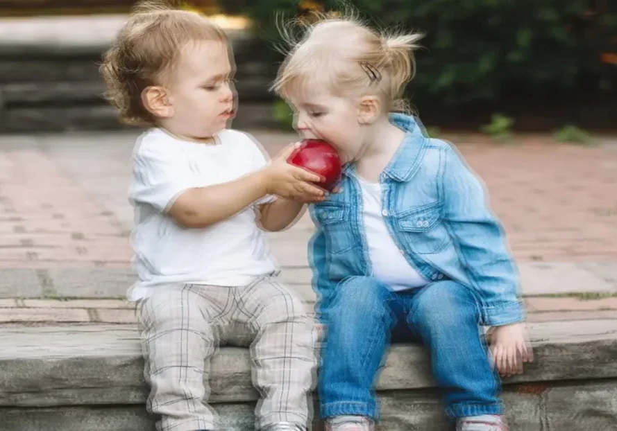Two toddlers sharing a red apple while sitting on a stone step outdoors.