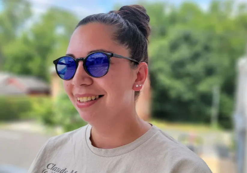 Smiling person wearing sunglasses with blue lenses outside on a sunny day.