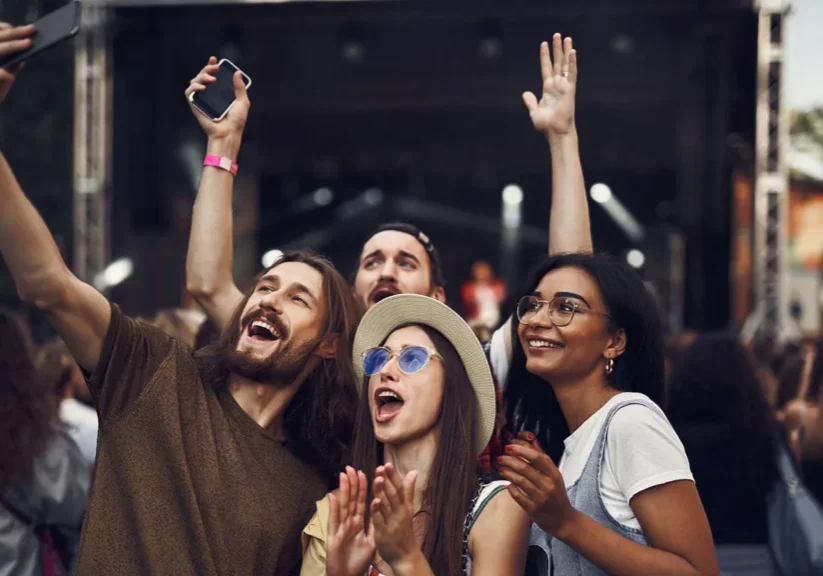 Group of friends enjoying a festival, taking selfies and cheering in front of a live stage.