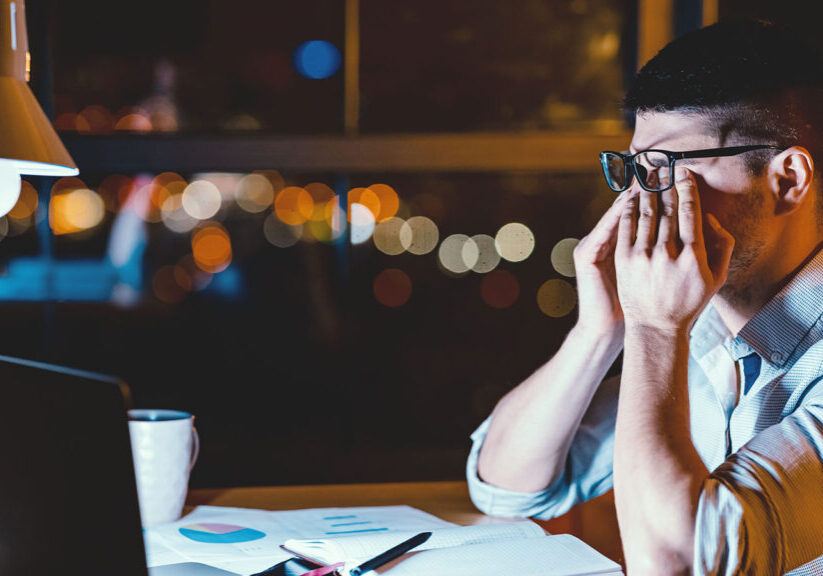 Man in glasses rubs eyes while working late on laptop, surrounded by papers and coffee, illuminated by desk lamp.