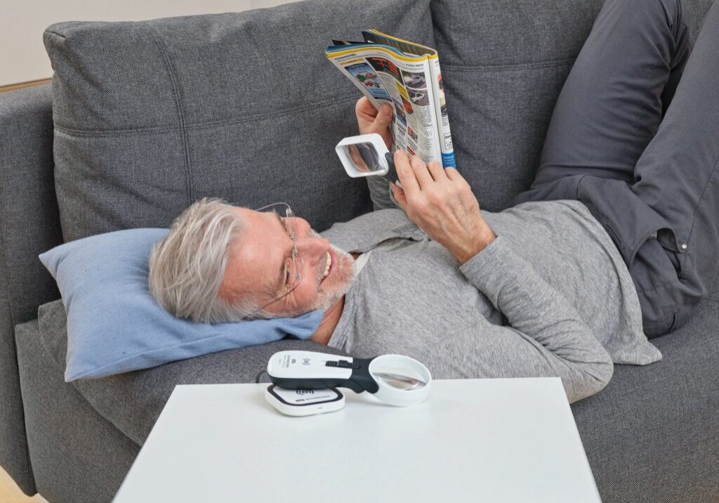 Man relaxing on a couch, reading a magazine with magnifier glasses, next to a table with magnifying tools.
