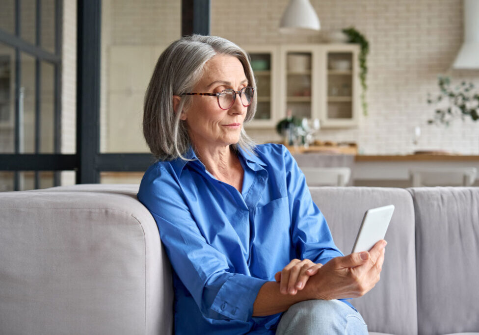 Older woman in blue shirt using smartphone on couch at home.
