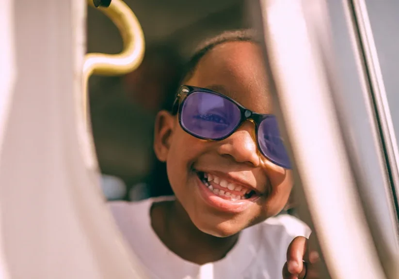 Smiling child with purple sunglasses, looking through a window, capturing joy and playfulness in the sunlight.