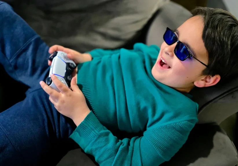 Child wearing sunglasses happily playing video games on a couch with a controller.