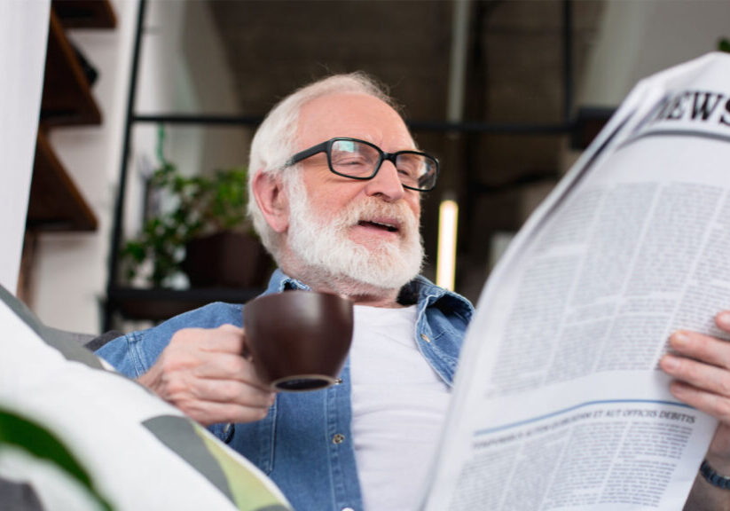 Elderly man enjoying a coffee while reading a newspaper at home, surrounded by greenery.