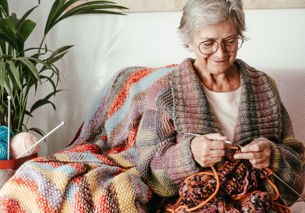 Elderly woman knitting colorful blanket, sitting on sofa, plants and yarn beside her. Cozy and creative atmosphere.