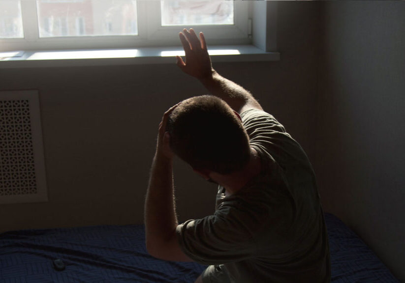 Person shielding eyes from sunlight near a window, sitting on a bed with a blue coverlet.