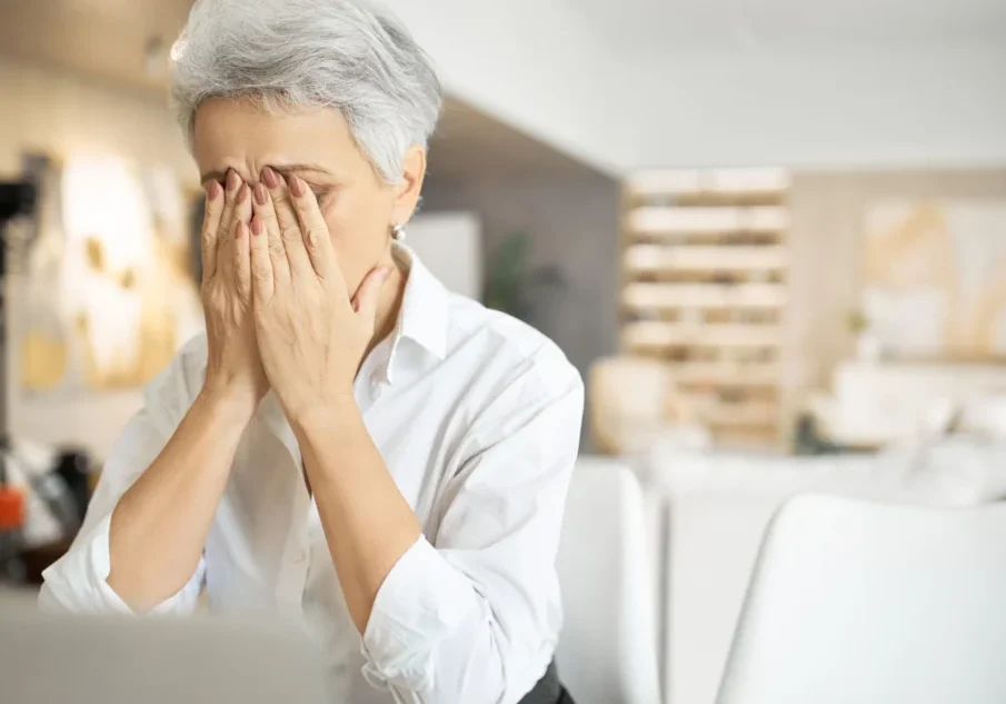 Stressed senior woman covering face with hands in bright office environment, expressing anxiety or fatigue.