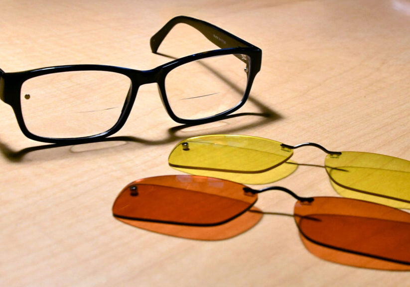 Black eyeglasses with interchangeable yellow and orange lenses on a wooden surface.