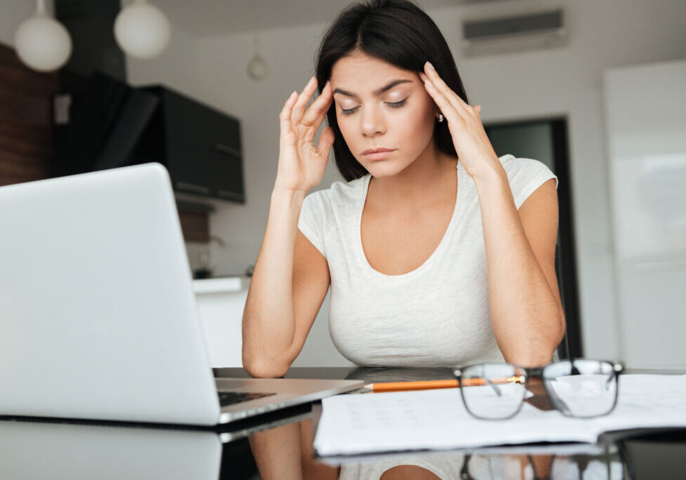 Woman feeling stressed at work, sitting at a desk with a laptop, holding her head in her hands.